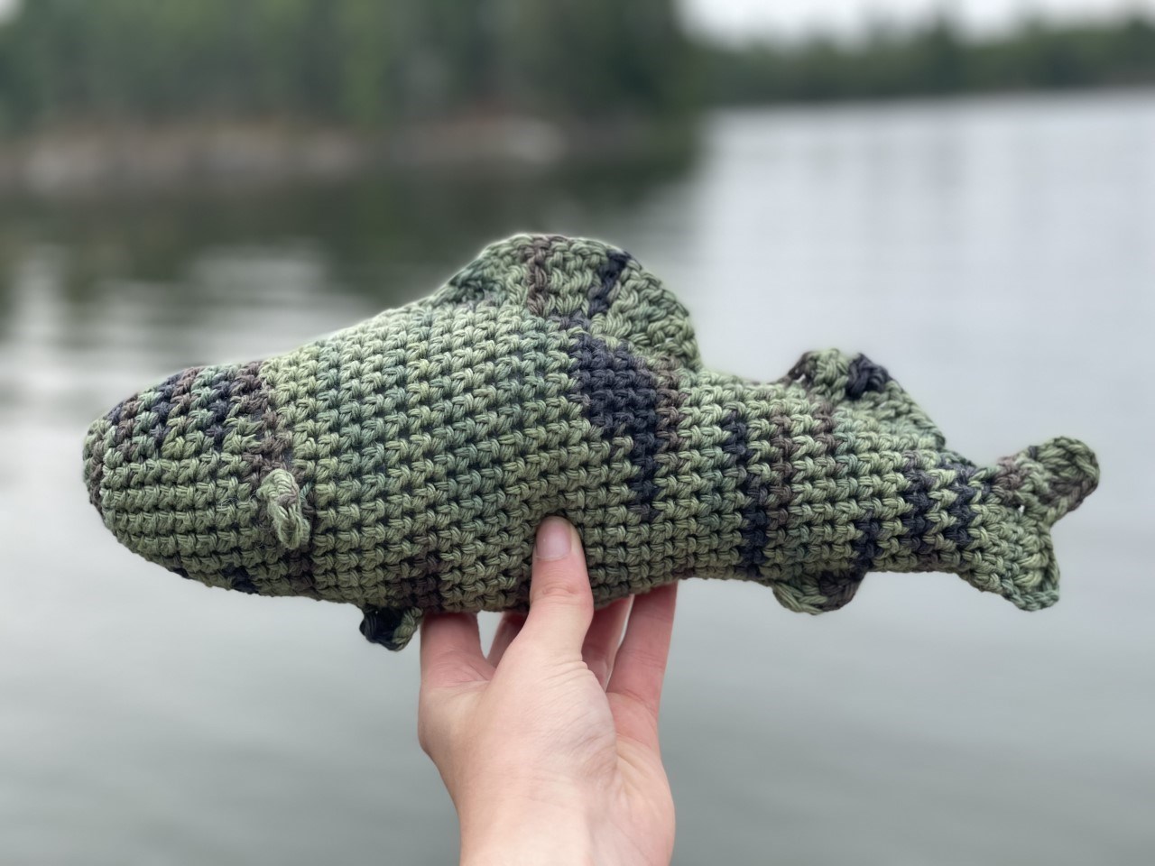 A crocheted fishing being held by a hand in front of a lake.