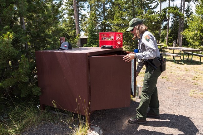 a woman in national park service uniform closes a brown bear resistant food locker in a campground