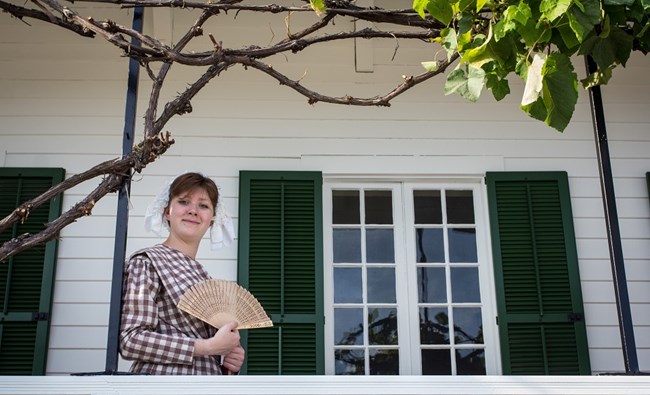 A young woman holding a fan stands on the veranda of the Chief Factor's House.