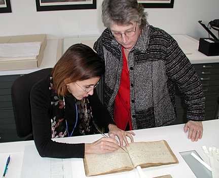 Two women stand over the old book. One places a long stick near the center fold. The other observes.