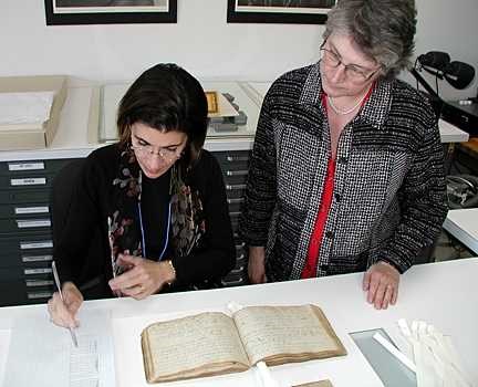 The two women stand over the old book. One makes notes while the other observes.