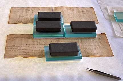 On a large table, old pages are covered in a glass square with a heavy looking weight on top.