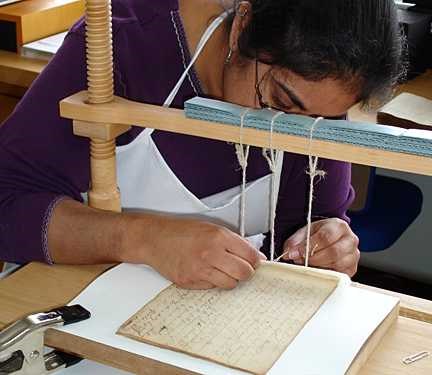 A wooden brace with 3 pieces of string dangling from it stands over the old book. A woman gently manipulates the strings.