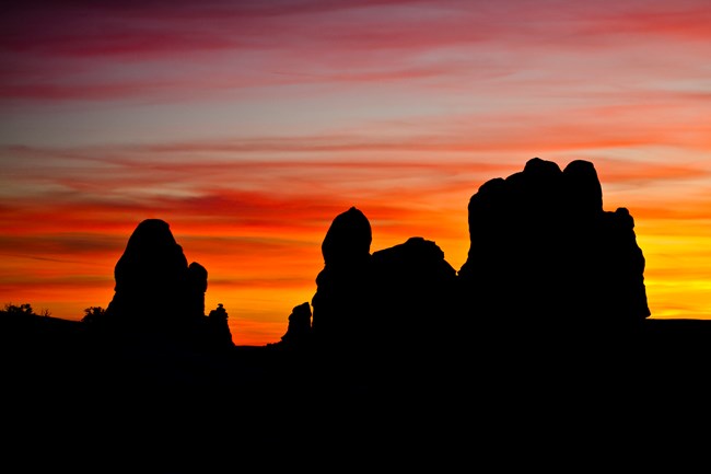 A yellow, orange, pink, and blue colored sunset behind black silhouettes of rock pillars.
