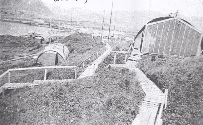 Grainy black and white photo of Quonset huts on a hill, with wooden boardwalks.