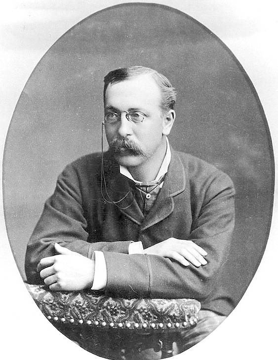 a man with glasses and a mustache who is leaning on the back of a chair
