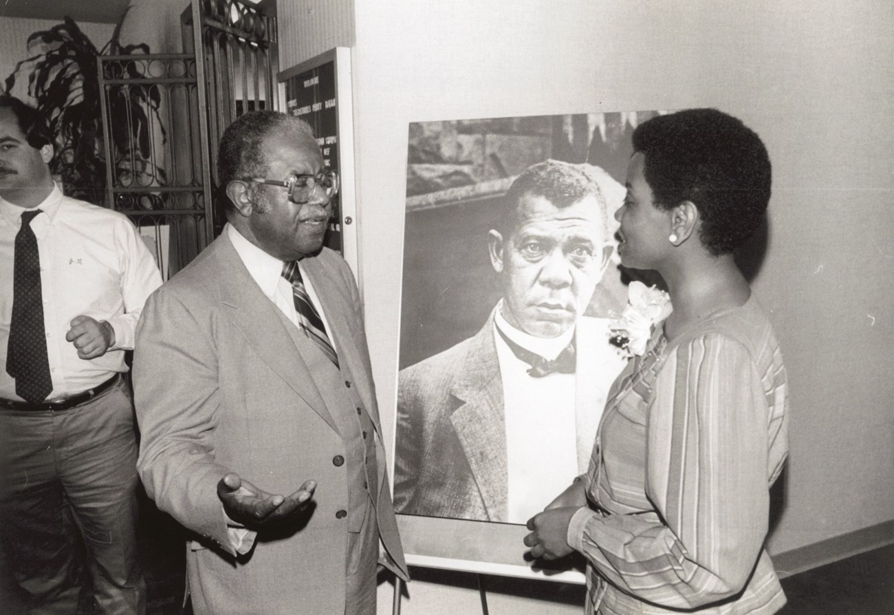 Superintendent Geraldine Bell and Reverend Ralph D. Abernathy A woman and a man stand chatting in front a photo of Booker T. Washington.