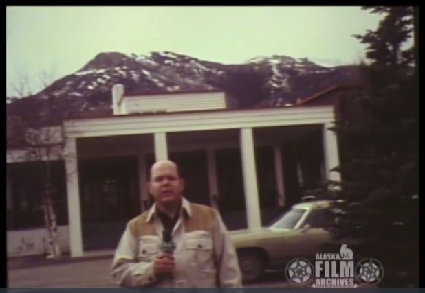 a reporter holding a microphone stands outside of a hotel near an old green car