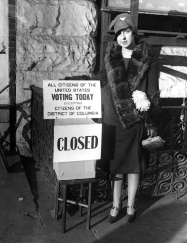 A black and white photo of a woman standing next to a sign that says: "all citizens of the united states voting today excepting citizens of the district of columbia"