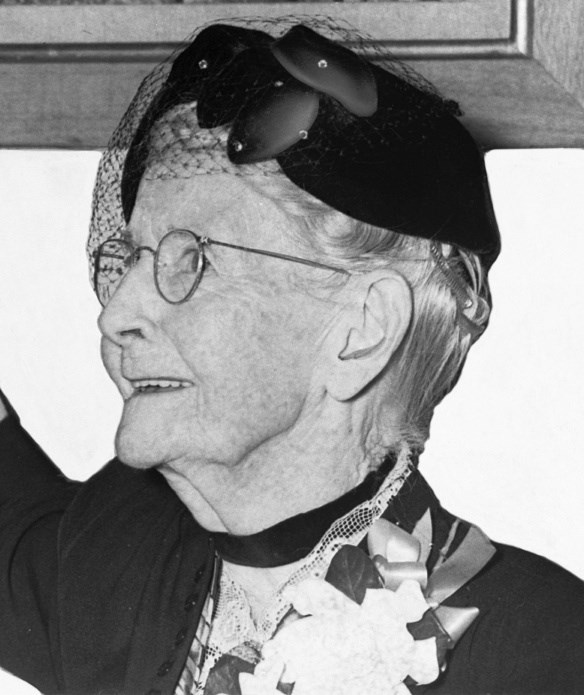 Grandma Moses woman wearing a dress, hat, and a glasses with a large white flower corsaige