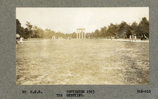 greeting Black and white photograph of large, flat open grassy area with walking paths on both sides, with some people using them. At the end are four large white pillars, with a top, connecting them all