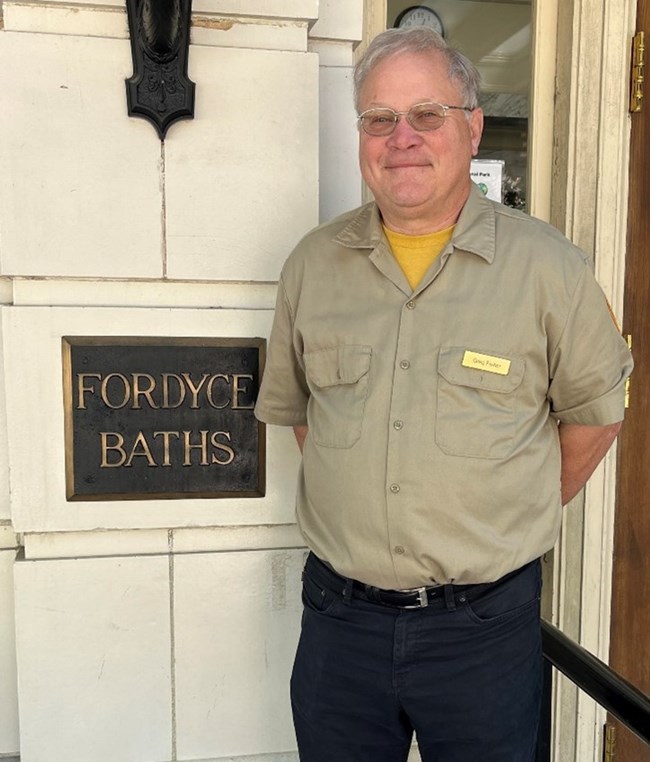 A man with gray hair and glasses wearing a beige button-down shirt poses next to a sign that reads, "Fordyce Baths."