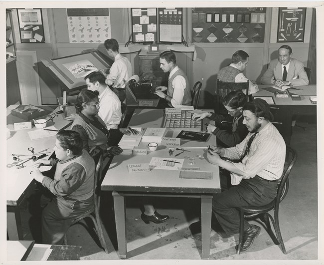 Black and white photo of adults sitting at tables with various tools and testing materials laid out in front of them. The group consists of 7 white men including the instructor and 1 Black woman.