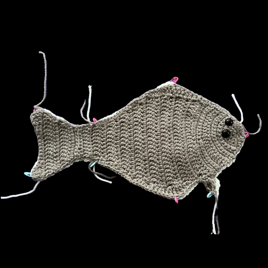 Two halves of a halibut fish that has been crocheted with yarn is held together by small pins.