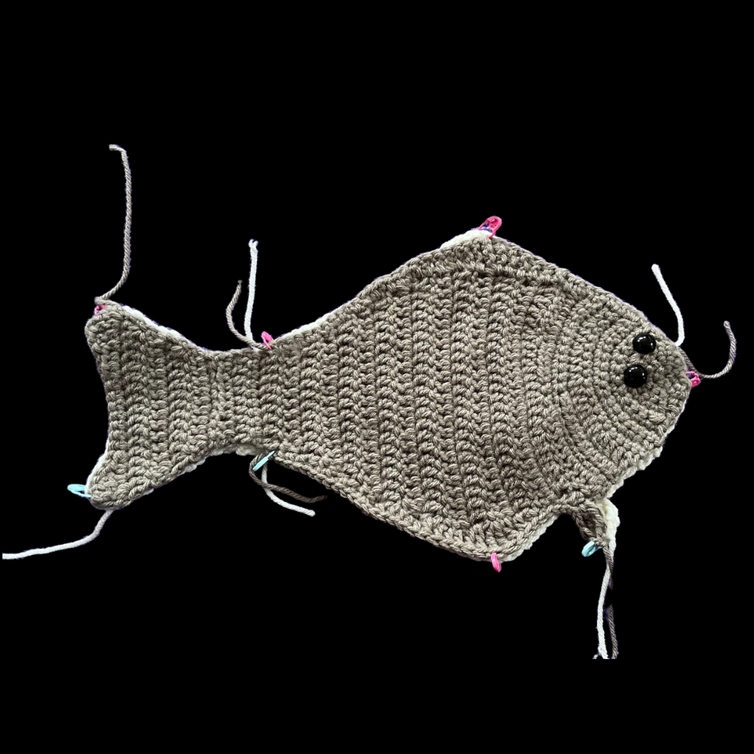 lt= Two halves of a halibut fish that has been crocheted with yarn is held together by small pins.