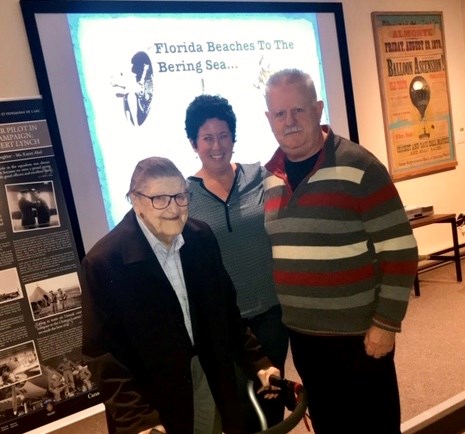 Harold Cross, Karen Abel, and Jon Ambler in 2018. Two men with white hair standing on either side of a woman with dark hair, in front of a screen.