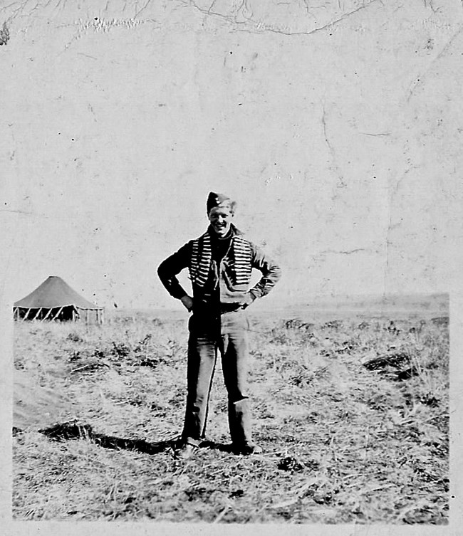Man in uniform with string of large 50 caliber bullets draped around his shoulders, with his hands on his hips, standing in a barren landscape.