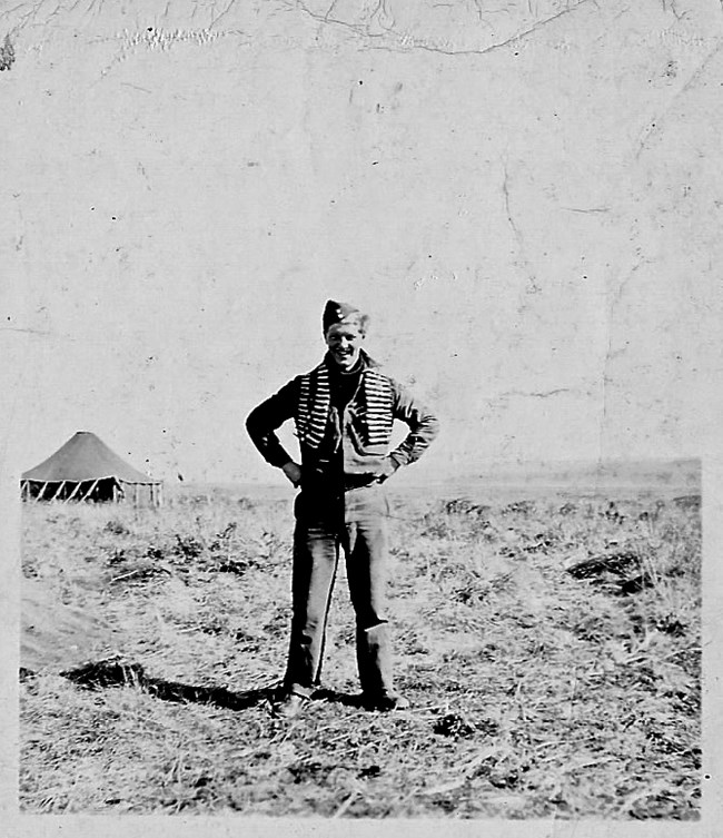 Harold Cross at Ft. Glenn, Umnak, 1942 Man in uniform with string of large 50 caliber bullets draped around his shoulders, with his hands on his hips, standing in a barren landscape.