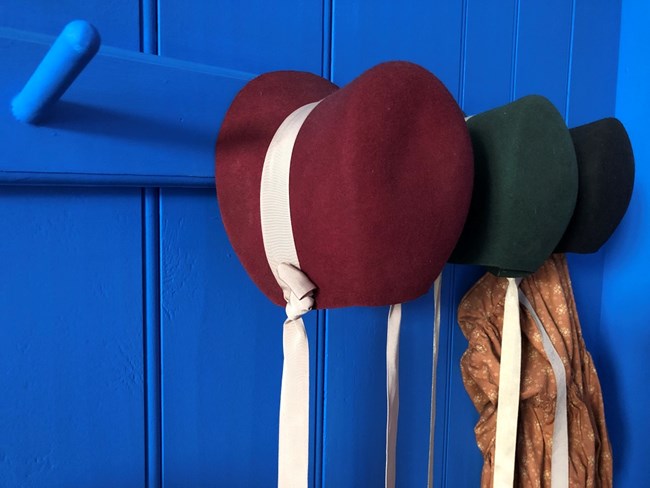 A bright blue painted wall with a series of pegs. One is red, one is green, and one is dark blue. All are decorated with simple ribbons. three of the pegs are felt girls' bonnet-style hats. They are
