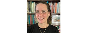 Woman with brown hair, glasses, and a black shirt smiling for the photo in front of a packed bookshelf.