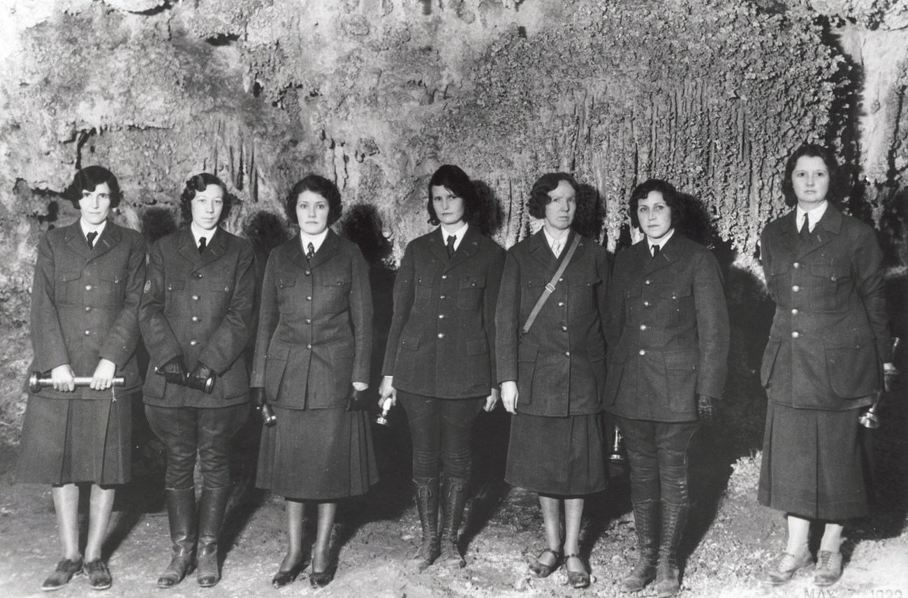 Seven women wearing NPS uniforms stand in a row in front of a rock wall.