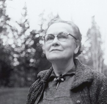 A woman wearing glasses and a coat stands in the forest looking to the left.