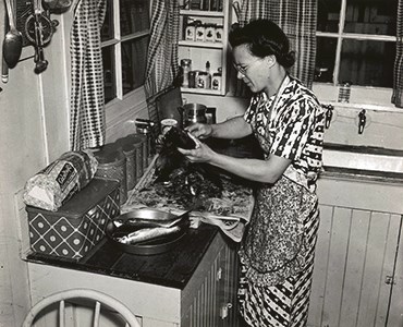 Woman at stove wearing an apron over her dress.  She is preparing some kind of meal in her kitchen.  The sink can be seen to the back right of the room.