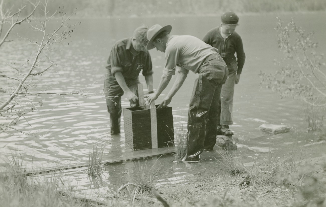 Three men, one in a ranger uniform, stand in a river over a large box.