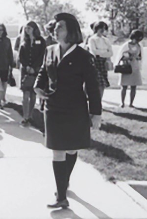 A woman wears a white shirt under a dark jacket. Her skirt falls just at her knee with dark socks that come up just under her knee. she stands with one leg crossed in front of the other while a group of people stand behind her.