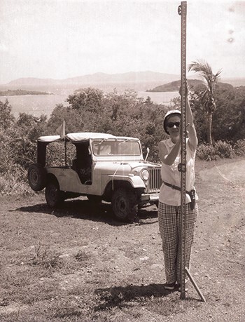 a woman wearing pants, sleeveless shirt, sunglasses and pith hat holds a long survey pole upright. A jeep can be seen to the back left of her.