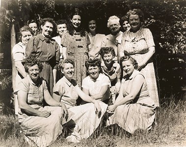 A group of women all wearing dresses poses in front of some trees the women in the front all kneel on the ground and they all smile at the camera.
