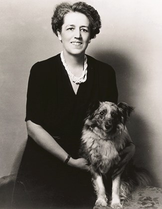 A woman wearing a dark dress and multiple strands of pearls sits next to a dog. The both face forward to the camera.