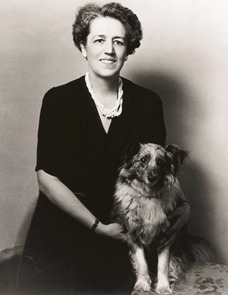 hfca_1645_cooper_gertrude_1940_u A woman wearing a dark dress and multiple strands of pearls sits next to a dog. The both face forward to the camera.