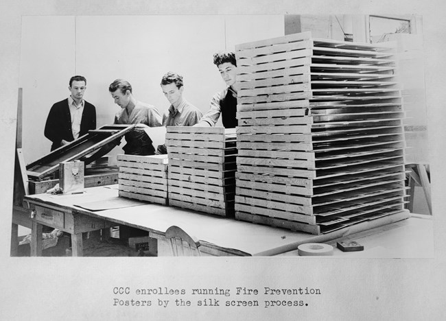 Four men work a silkscreen press amid drying racks