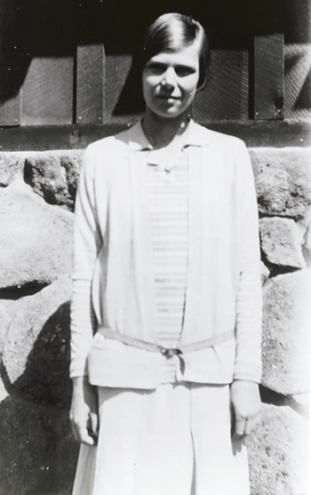 Ruth Ashton wears a long skirt, a collared blouse and belted cardigan stands facing the camera.