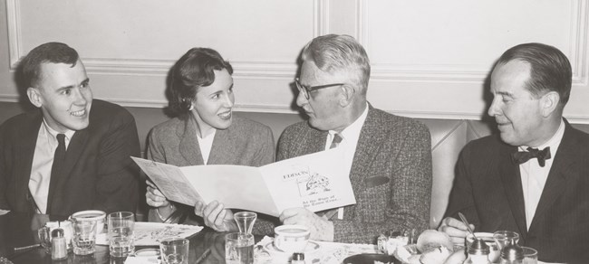 Three men and a woman sit at a table looking at a brochure