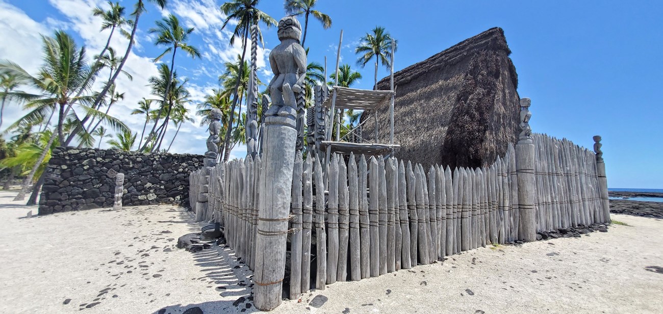 Hale o Keawe from corner fencepost Surrounded by a wooden palisade, thatched Hale o Keawe stands amid carved kiʻi and lele (offering platform). The edge of the Great Wall is visible behind Hale o Keawe.