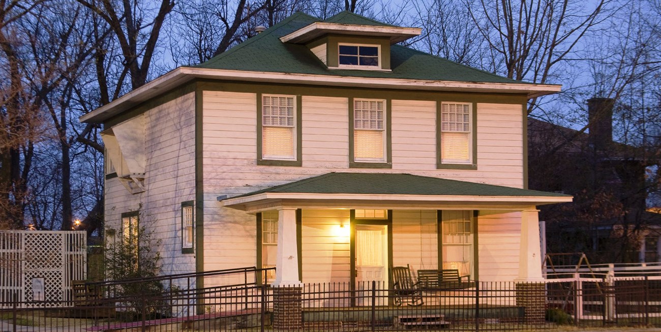 White two story house lit at dusk with green trim and trees behind.