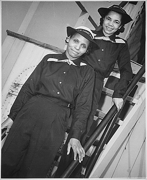 Two African American women in Coast Guard uniforms smile at the camera. They stand on a set of steep stairs.