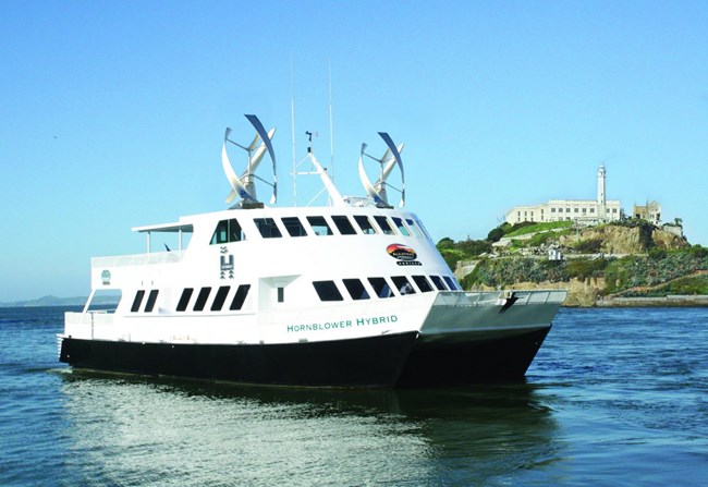 A ferry in front of Alcatraz Island. The ferry says "Hornblower Hybrid" on the side, and has two large wind turbines on top.