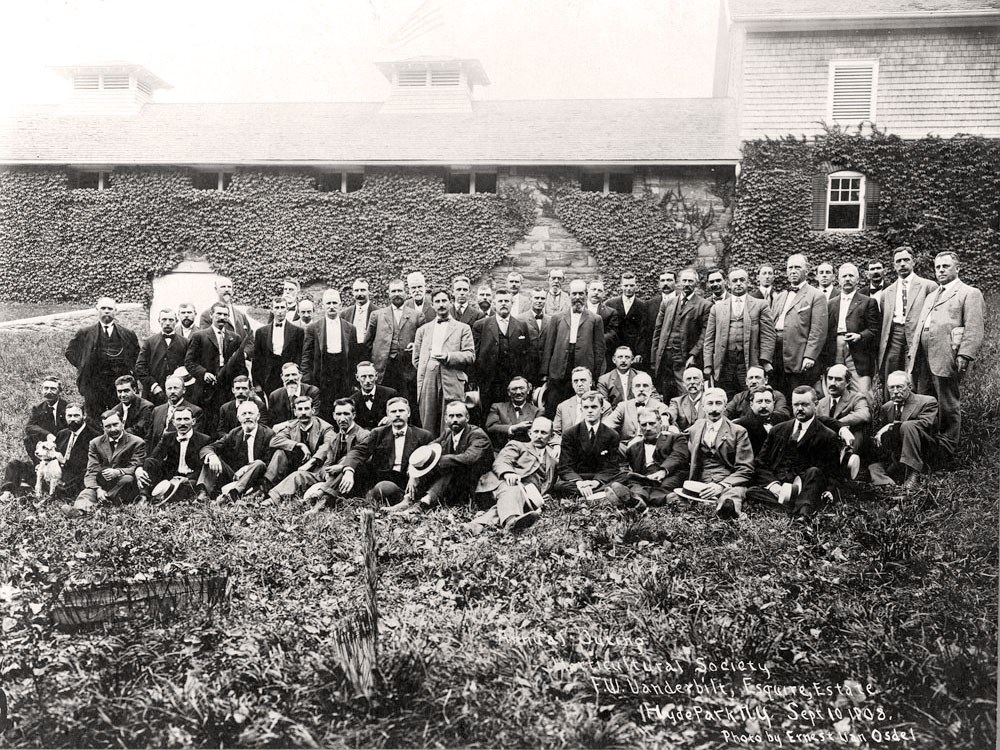 A large group of men seated on a lawn before a barn complex.