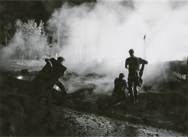 hotel fire 1 A black and white photo of three people facing toward a smoky area, holding various large objects. Smoke clouds the view of trees in the distance.