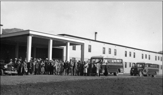 A black and white photo of a long building with two rows of many windows down the length of the building. A crowd of people stands near the entrance. Three vehicles are parked off to the side.
