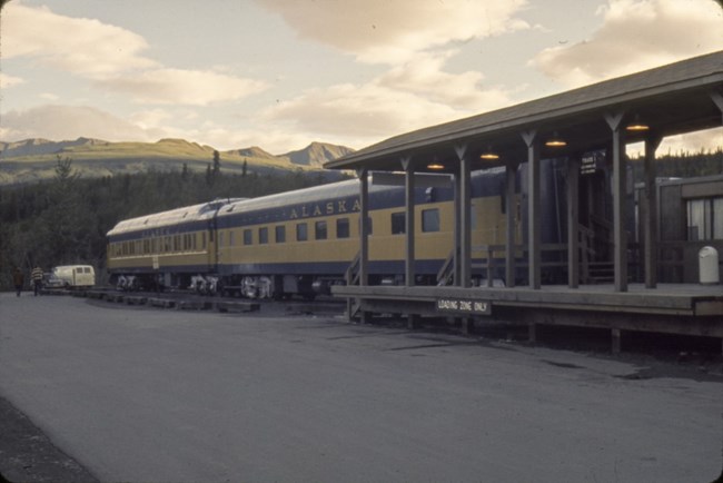 hotel fire 3 Two blue and yellow train cars with the word Alaska on the side sit next to a wooden platform. Trees and mountains are visible in the distance.