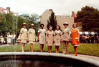 Seven women stand in a line on a sidewalk.  Six of them wear tan dresses and the one on the far right wears an orange smock.