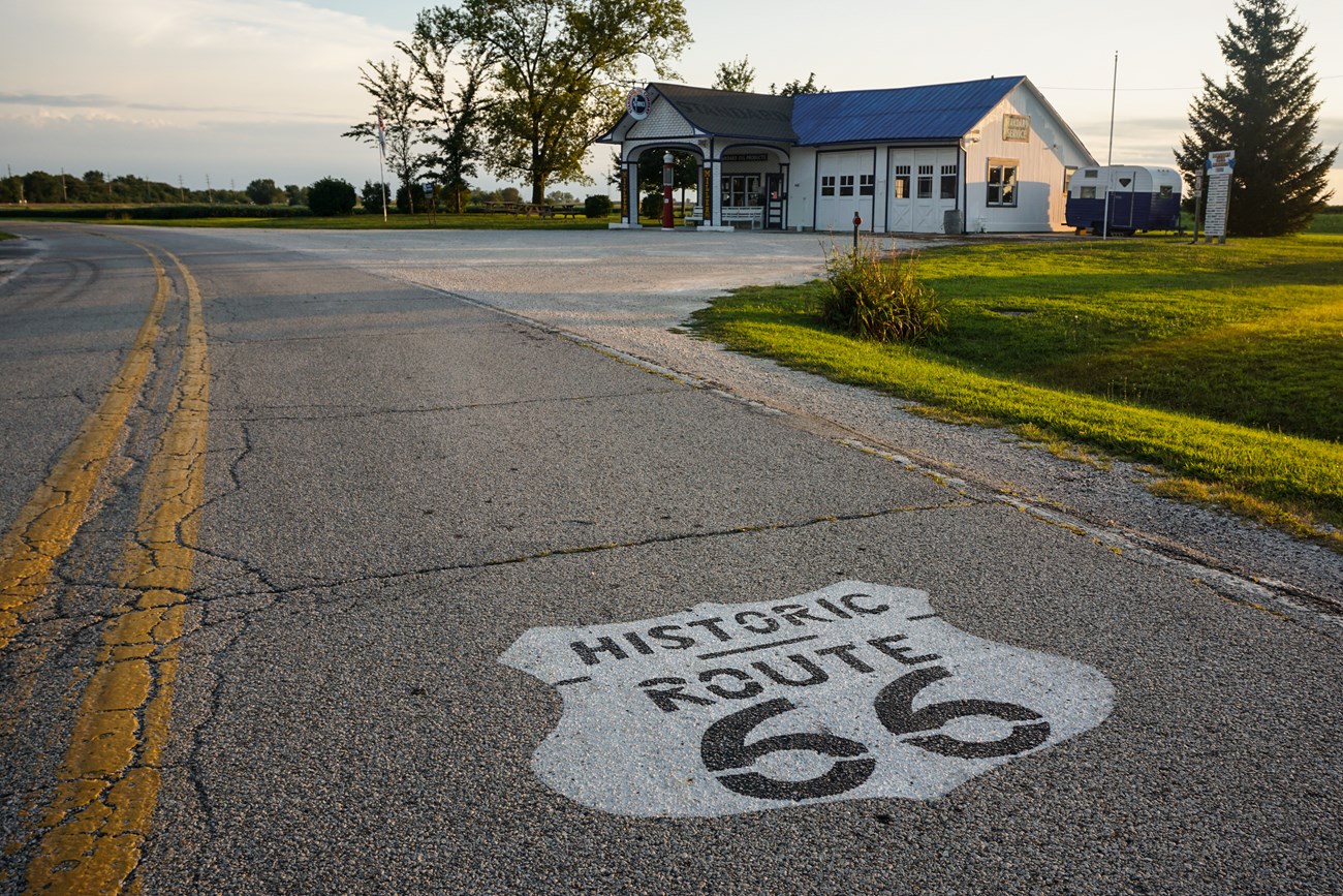 A paved road with a white Route 66 emblem painted on leads up to a white old-fashioned gas station with a green lawn and trees behind.