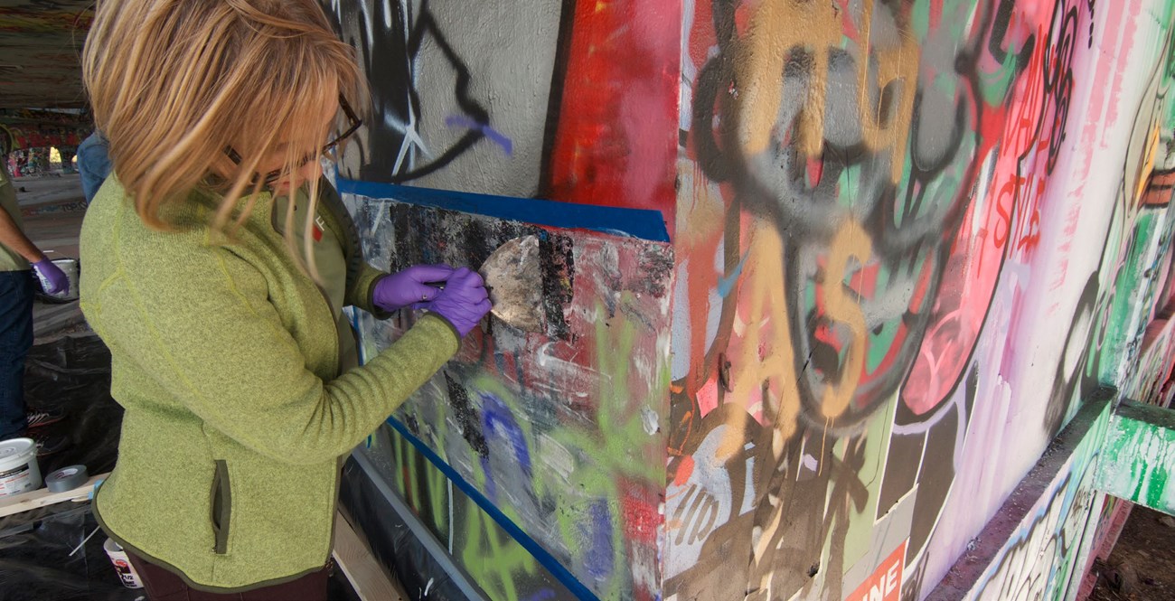 A preservationist is stripping the first layer of paint from a concrete structure that is covered in graffiti