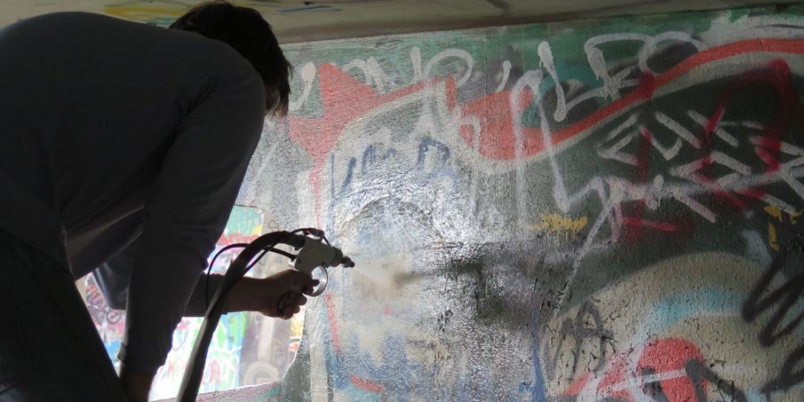 Preservationist using an air spray gun on the surface of concrete covered in graffiti Man using Air Spray Gun on Graffitied concrete wall