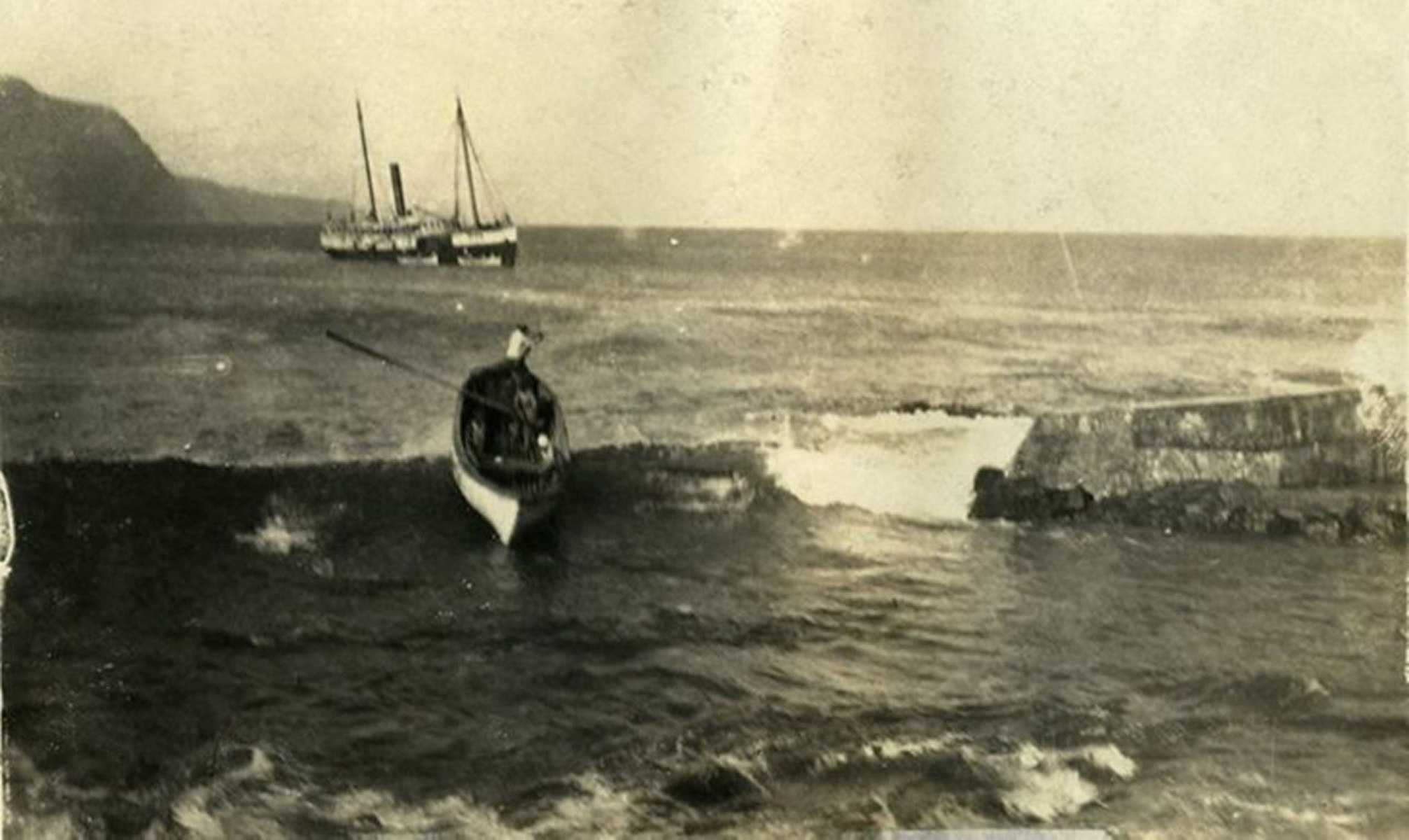 Outdoors; sepia photo of person sitting on a break wall with ocean and boat behind.