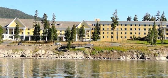 Photo taken from the lake of a large yellow hotel and road between the hotel and the water. A wooden viewing platform rests on top of a sandy bluff close to the edge. Water is in the foreground.